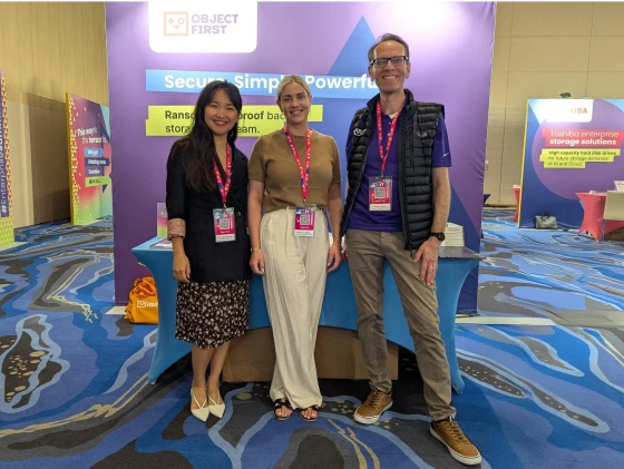 Three Object First team members posing and smiling in front of an “Object First” exhibition booth at a tech event.