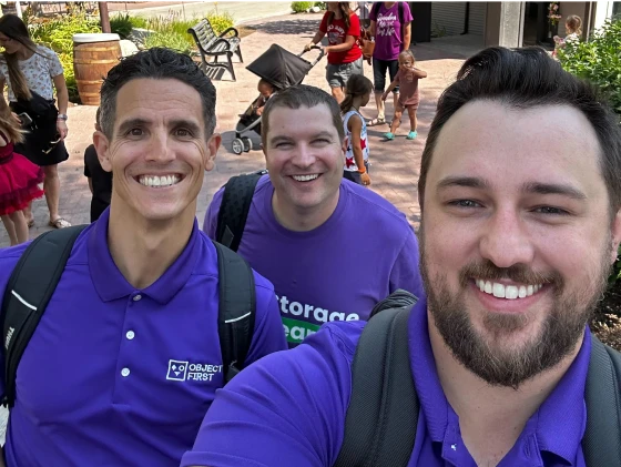 Three men taking a selfie outdoors, all smiling, wearing purple shirts, and carrying backpacks.